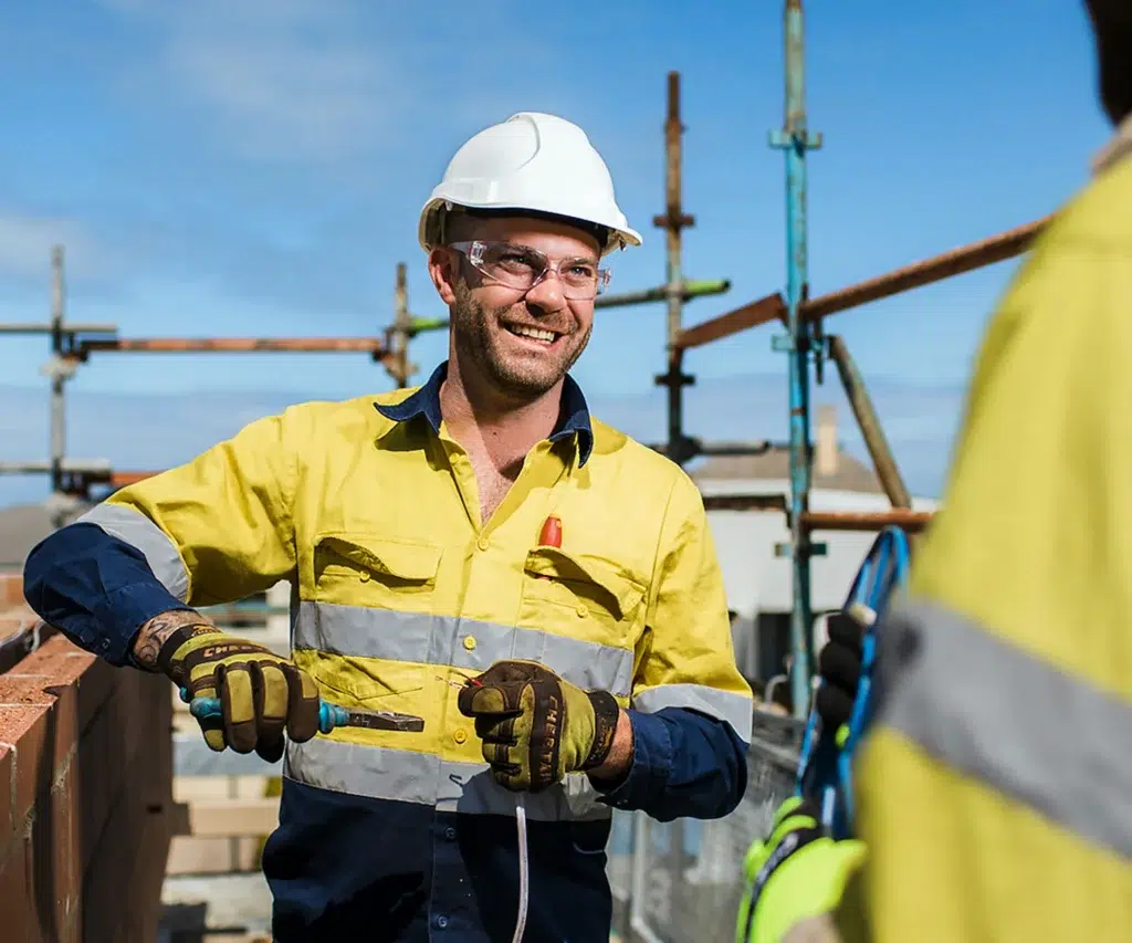 A smiling construction worker in a yellow high-visibility shirt, gloves, and a white hard hat holds tools at a worksite with scaffolding behind—showcasing teamwork and skill like no other. Another worker is partially visible in the foreground.