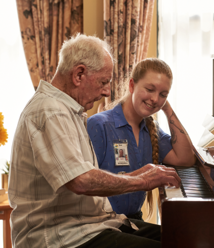 An elderly man plays the piano while a young woman in a blue shirt with a Perth City Dental name badge sits beside him, smiling and watching. Sunlight streams through curtains in the background as they enjoy the moment indoors.