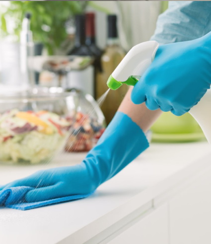 A person wearing blue gloves sprays cleaner and wipes a white kitchen worktop; a bowl of salad and other kitchen items are visible in the background, reflecting the high hygiene standards embraced by Perth City Dental.
