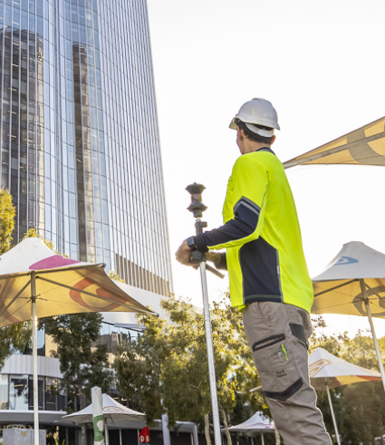 A construction worker in a high-visibility shirt and hard hat uses surveying equipment outdoors near Perth City Dental, with tall parasols and a modern glass building in the background.