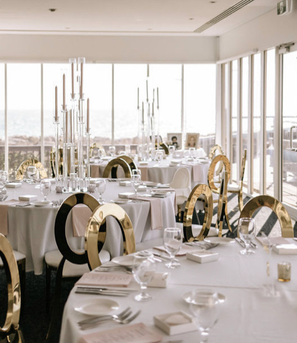 Elegant banqueting hall with round tables covered in white linen, gold and black chairs, tall glass candleholders, and large windows letting in natural light with a scenic Perth view—perfect for gatherings near City Dental.