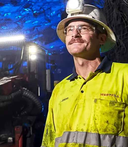 A male miner wearing glasses, a helmet with a headtorch, and a yellow safety shirt stands underground with mining equipment and blue lighting in the background, representing Westfill Group’s commitment to safe mining practices.