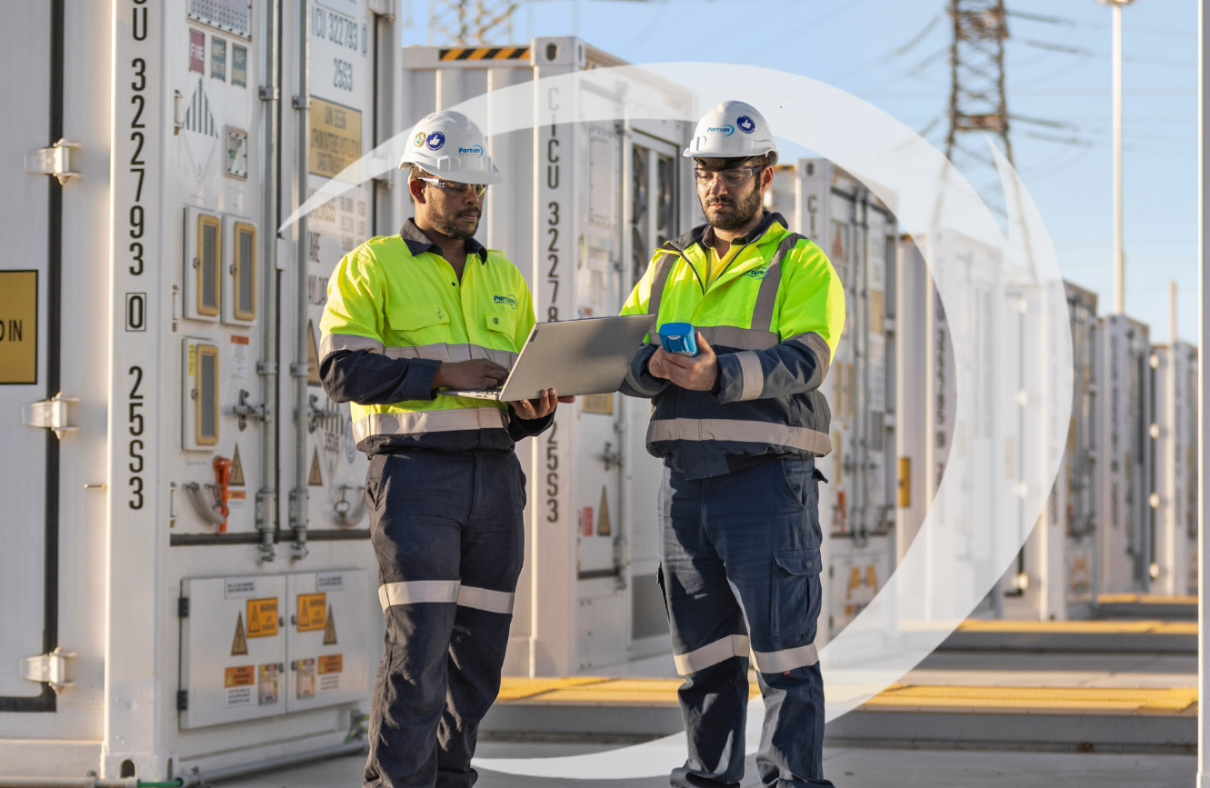 Two workers in high-visibility uniforms and helmets stand outside at an industrial facility, looking at a laptop. Shipping containers with control panels line the area. It’s sunny, power lines are visible, and the site reflects Partum Engineering’s expertise.