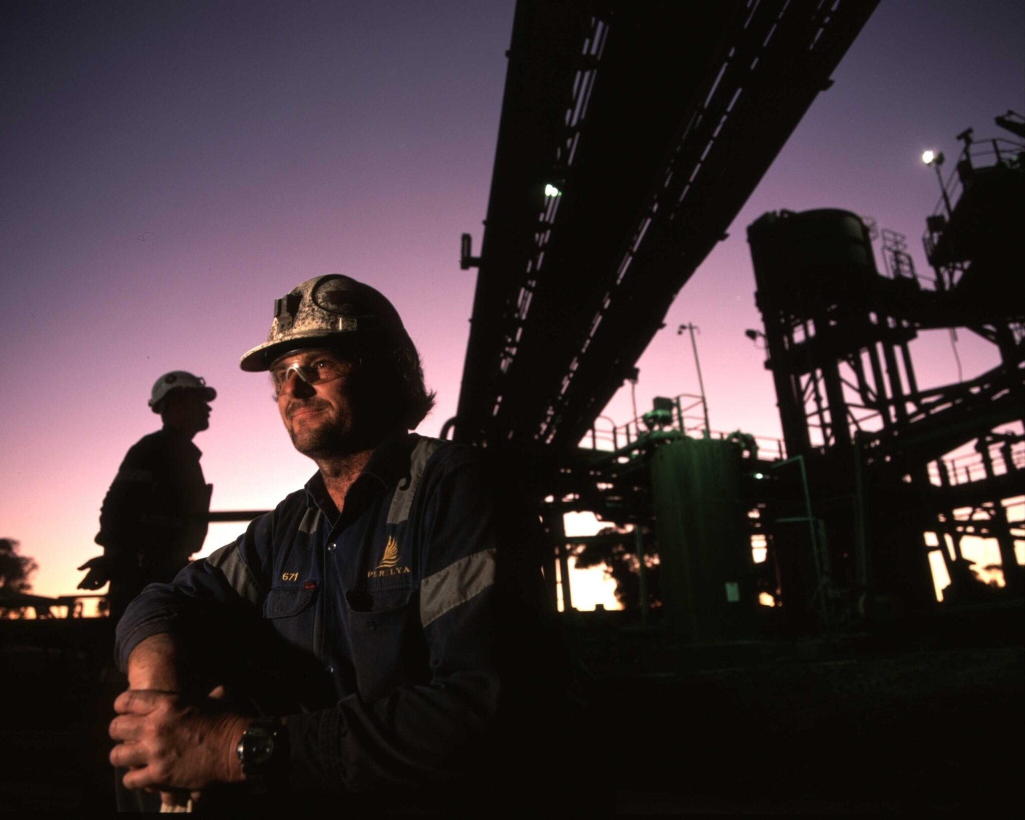 Two workers wearing helmets and safety glasses are at an industrial site at dusk. One worker is in the foreground with arms resting on his knees, whilst pipes and structures are silhouetted against the purple sky.